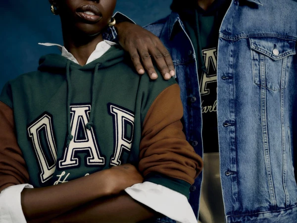 Close-up of two individuals posing together wearing Gap brand logo hoodies and a denim jacket against a deep blue background.