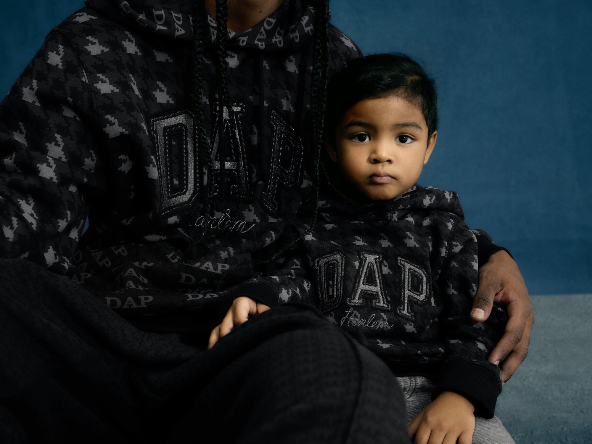 Close-up shot of an adult and child wearing matching patterned hoodies with 'DAP Harlem' text, set against a simple blue backdrop.