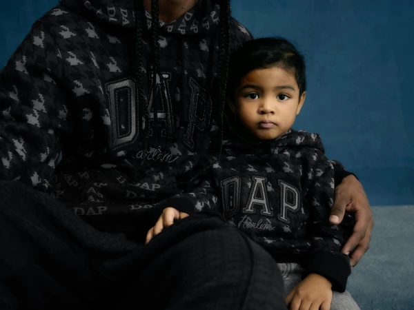 Close-up shot of an adult and child wearing matching patterned hoodies with 'DAP Harlem' text, set against a simple blue backdrop.