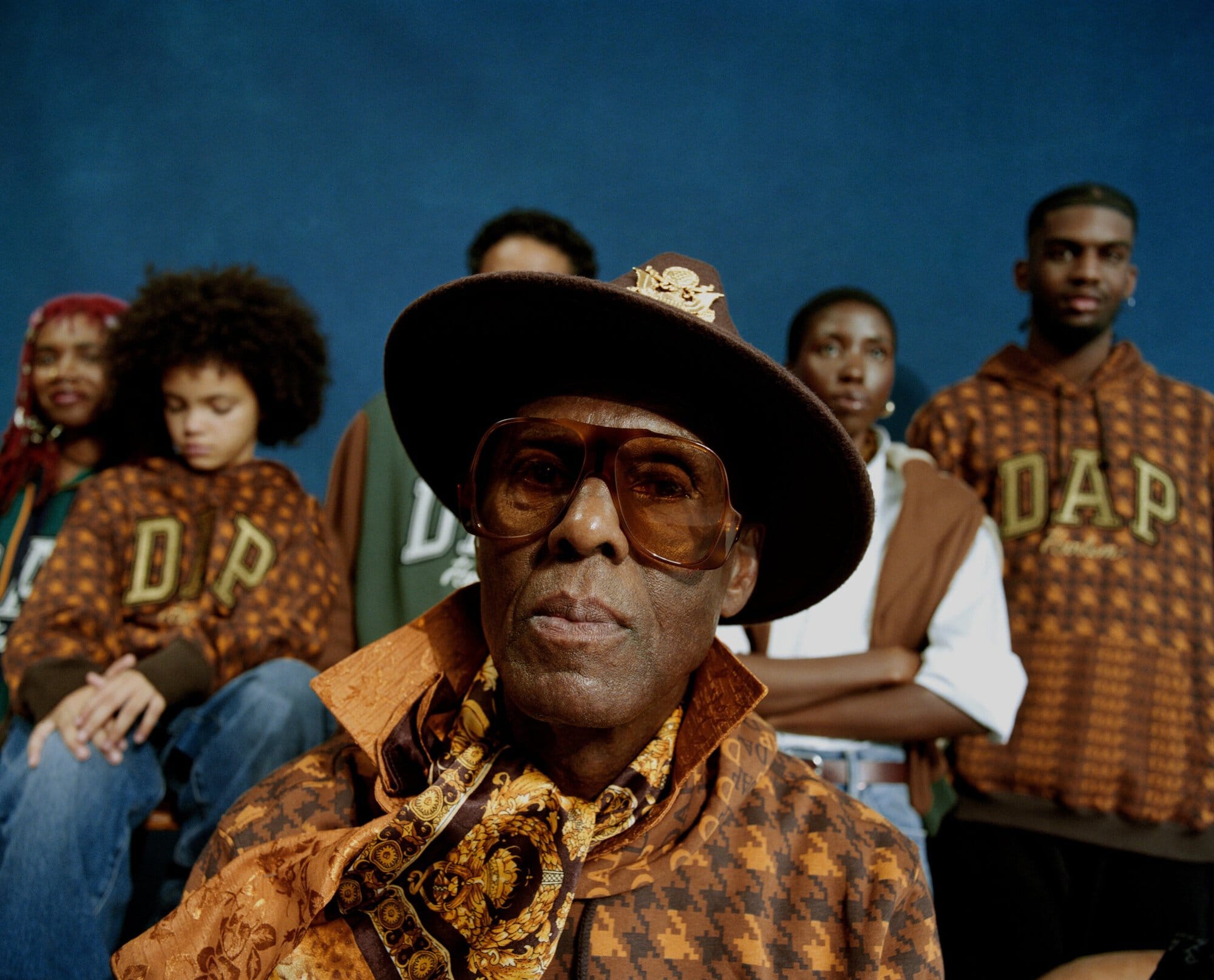 Group portrait featuring a mature African American man in a brown hat and patterned jacket, alongside others in matching hoodies against a blue backdrop.