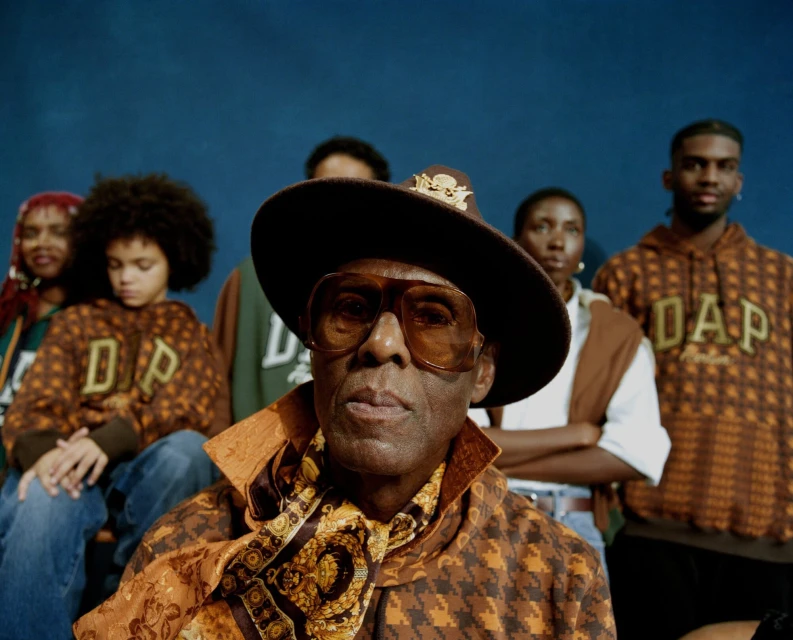 Group portrait featuring a mature African American man in a brown hat and patterned jacket, alongside others in matching hoodies against a blue backdrop.