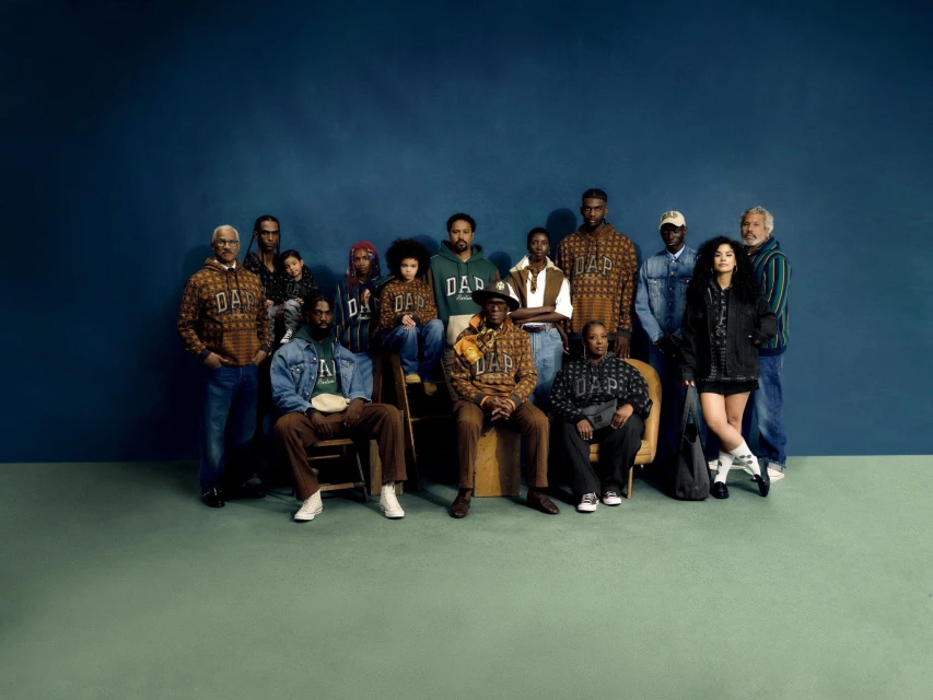 Group portrait of 14 people in casual fashion, modeling streetwear style GAP hoodies and denim in front of a muted teal background.