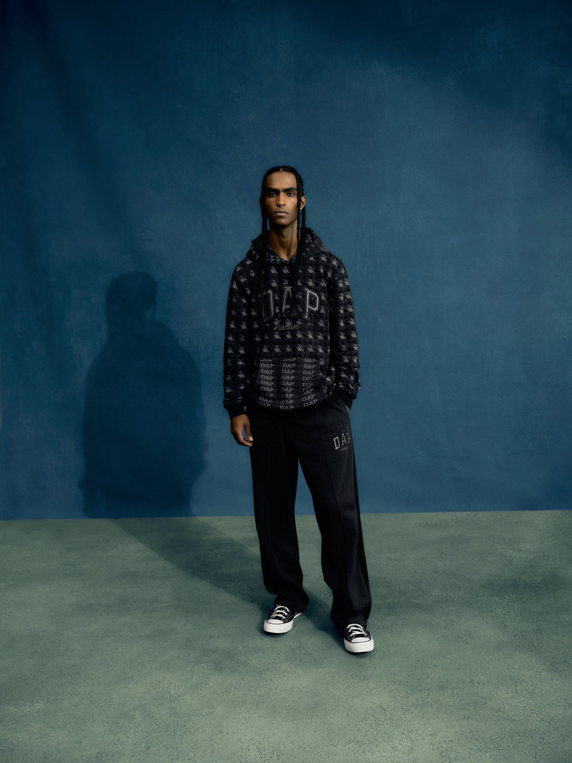 Man in a patterned GAP hoodie and sweatpants stands in front of a textured teal background in a full-length studio shot.