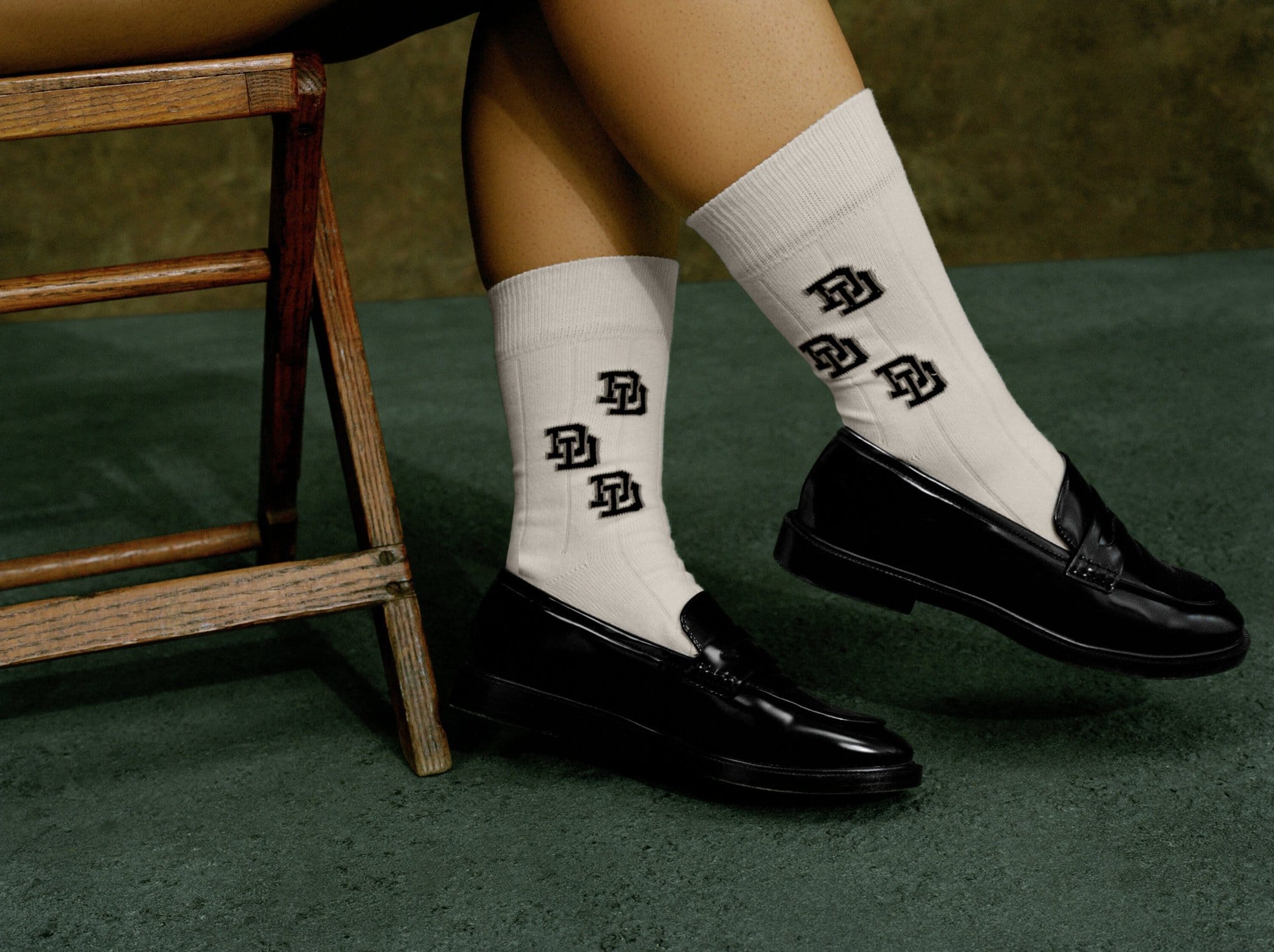 Close-up of legs wearing white socks with black logos and shiny black penny loafers, seated on a wood chair, green carpet.