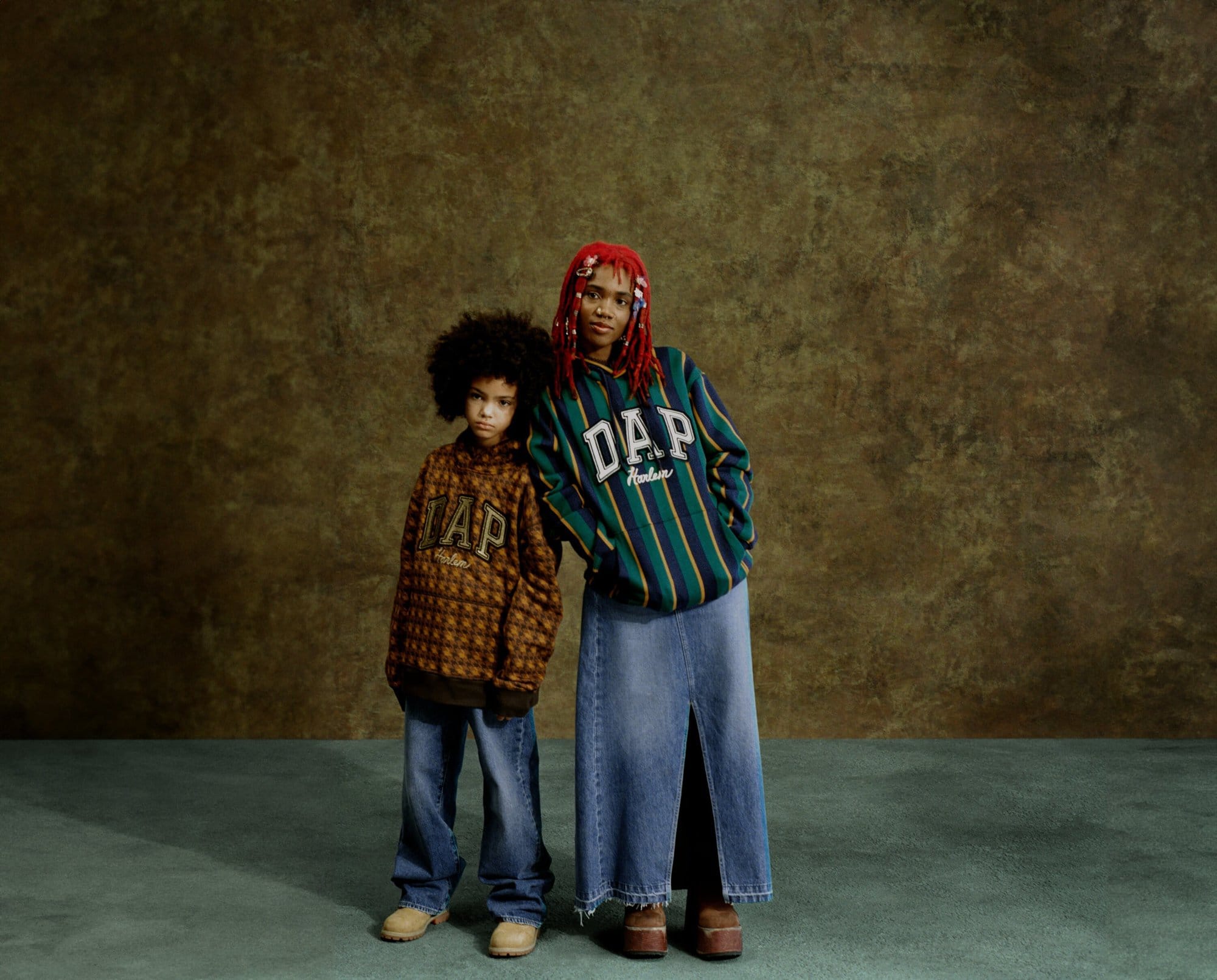 Two people, a young child and an adult, stand in front of a textured olive backdrop wearing GAP-branded clothing, shot in studio lighting.