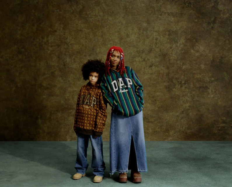 Two people, a young child and an adult, stand in front of a textured olive backdrop wearing GAP-branded clothing, shot in studio lighting.