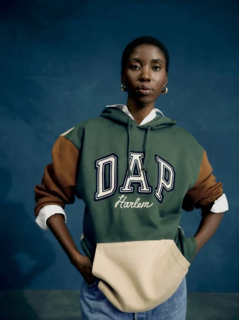A young Black woman poses indoors against a dark teal background, wearing a green, brown, and beige Gap hoodie with light blue jeans.