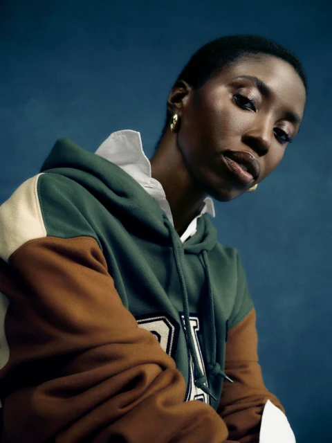 Portrait of a young Black woman in a color-blocked hoodie and gold earrings posing in front of a deep blue background.