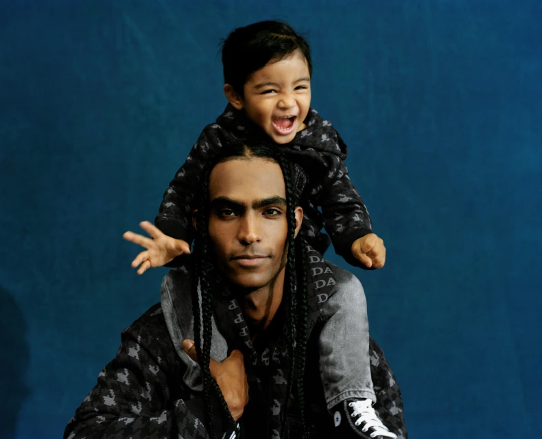 Adult male carrying toddler on shoulders, both in patterned sweatshirts against a dark blue backdrop. Smiling toddler's arm raised.