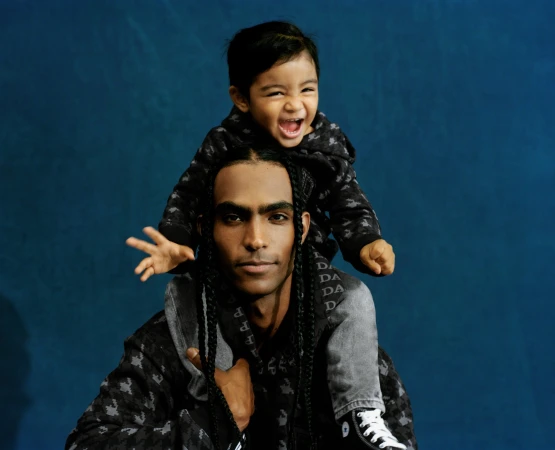 Adult male carrying toddler on shoulders, both in patterned sweatshirts against a dark blue backdrop. Smiling toddler's arm raised.