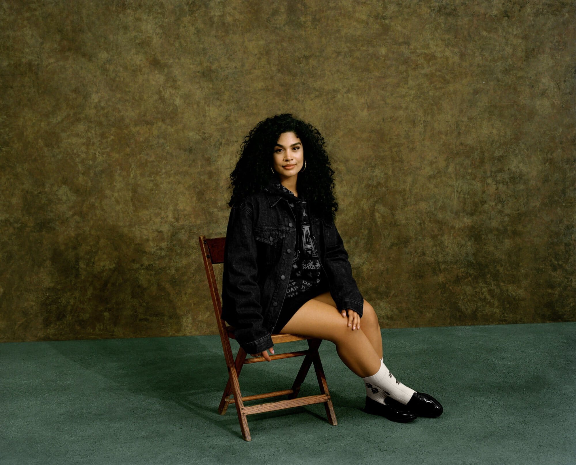A woman with curly hair sits on a wooden chair in a photo studio setting, wearing a black denim jacket and black shoes against a textured backdrop.