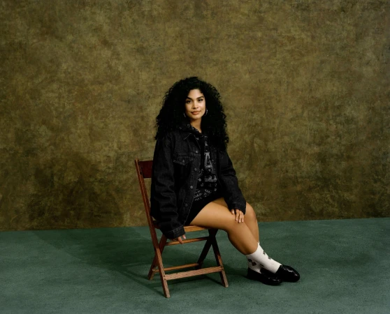 A woman with curly hair sits on a wooden chair in a photo studio setting, wearing a black denim jacket and black shoes against a textured backdrop.