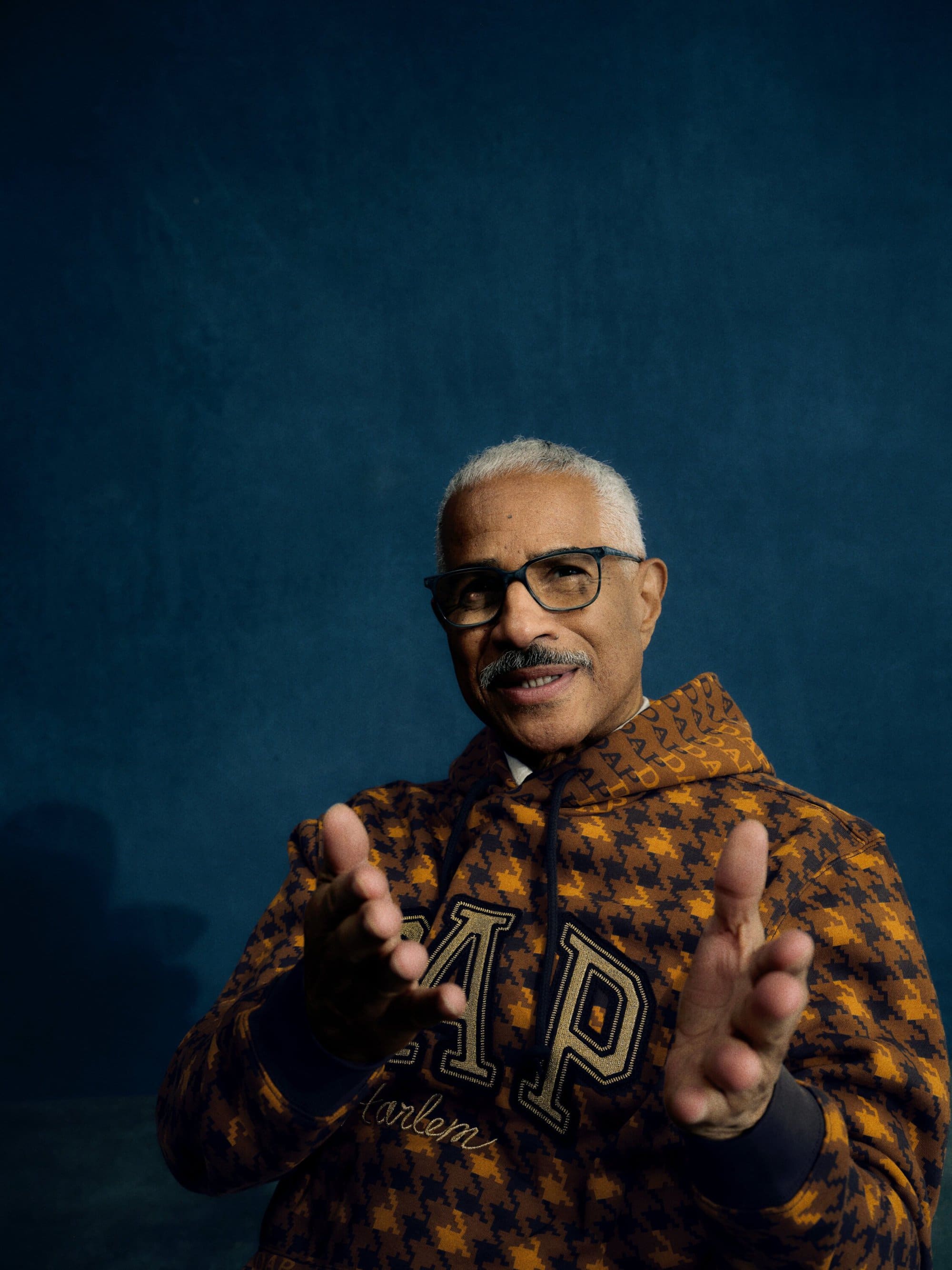 Portrait of a mature man wearing a GAP hoodie and glasses, with a mustache, clapping his hands in a photo studio against a blue backdrop.