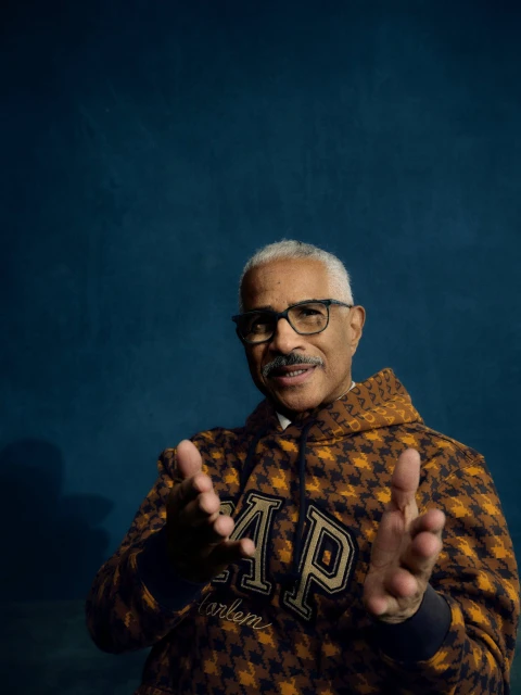 Portrait of a mature man wearing a GAP hoodie and glasses, with a mustache, clapping his hands in a photo studio against a blue backdrop.