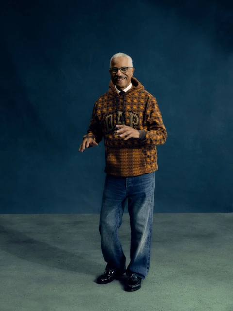 Older adult male in a patterned Gap hoodie, blue jeans, and black loafers posing against a dark blue studio backdrop with concrete flooring.