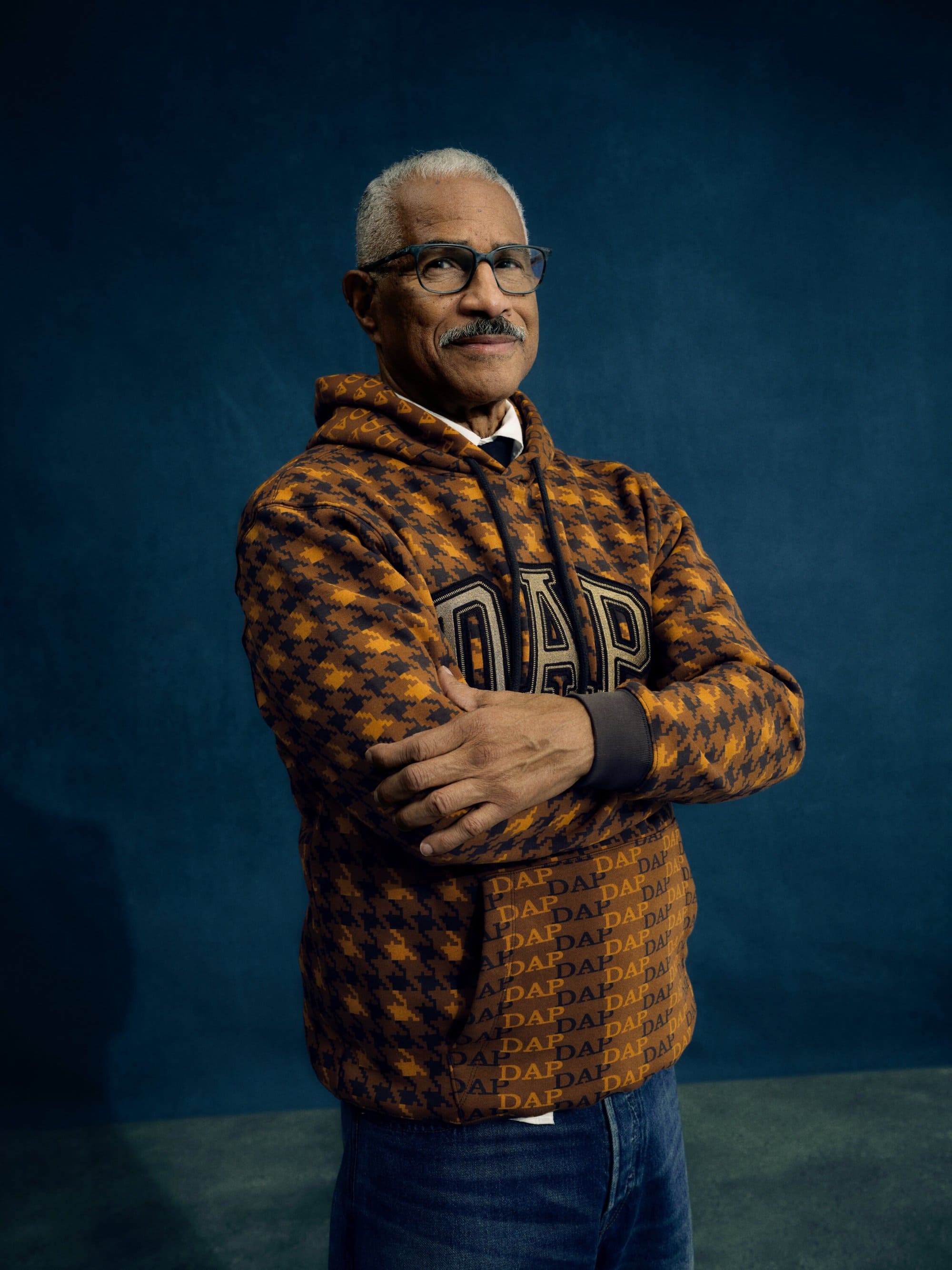 Portrait of an adult man with grey hair and a mustache, wearing glasses and a printed hoodie, posing against a dark blue background.