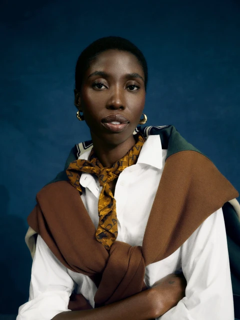 Dark-skinned adult female poses in a white shirt, with a brown scarf tied at the neck, against a dark blue backdrop in a medium shot.