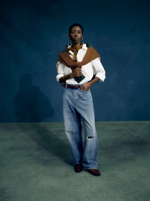 Full-length portrait of an adult woman in oversized denim jeans, a white button-down shirt, and brown loafers against a dark teal backdrop.
