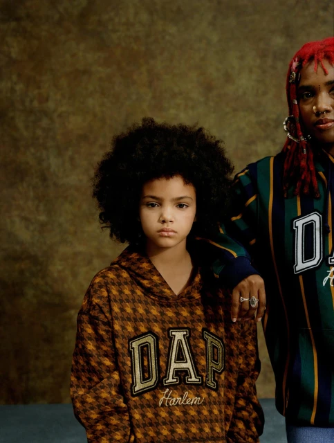 Two models wearing DAP hoodies pose against a textured brown backdrop, in a fashion-forward studio shot, showcasing urban youth style.