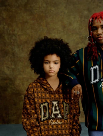 Two models wearing DAP hoodies pose against a textured brown backdrop, in a fashion-forward studio shot, showcasing urban youth style.