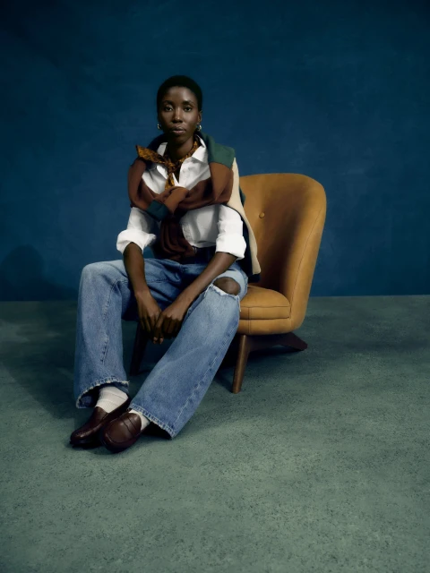 A woman with dark hair sits in a yellow chair, wearing a white shirt, ripped jeans, and loafers against a dark teal background.