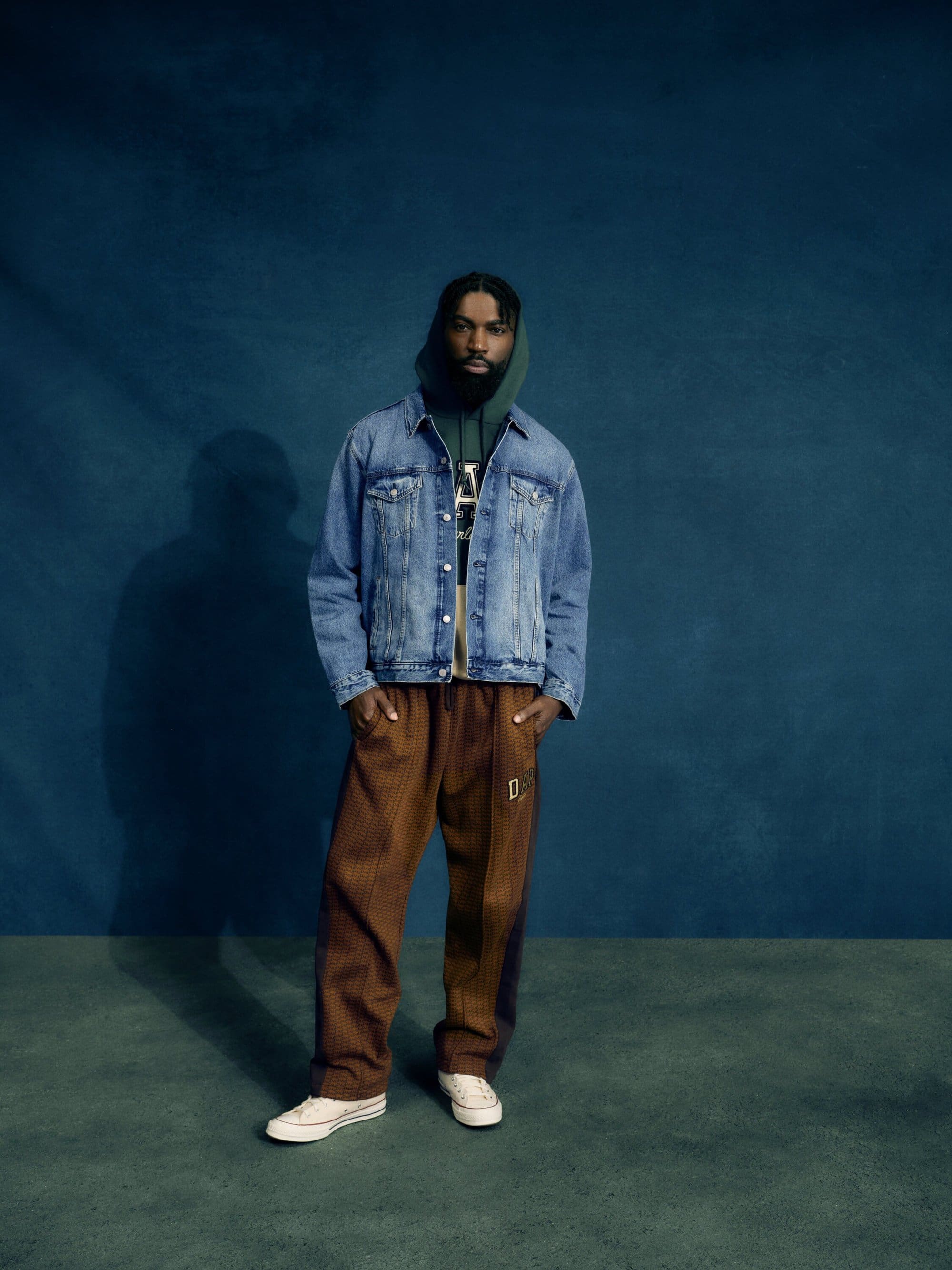Full-length studio shot of a Black male model wearing a denim jacket, green hoodie, brown pants, and white sneakers against a blue backdrop.