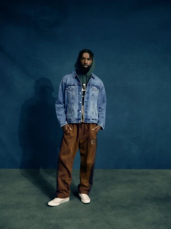 Full-length studio shot of a Black male model wearing a denim jacket, green hoodie, brown pants, and white sneakers against a blue backdrop.