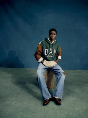 A Black woman poses in a studio, wearing a Gap hoodie and jeans, seated on a wooden block against a teal backdrop. The photo captures a casual, fashionable look.