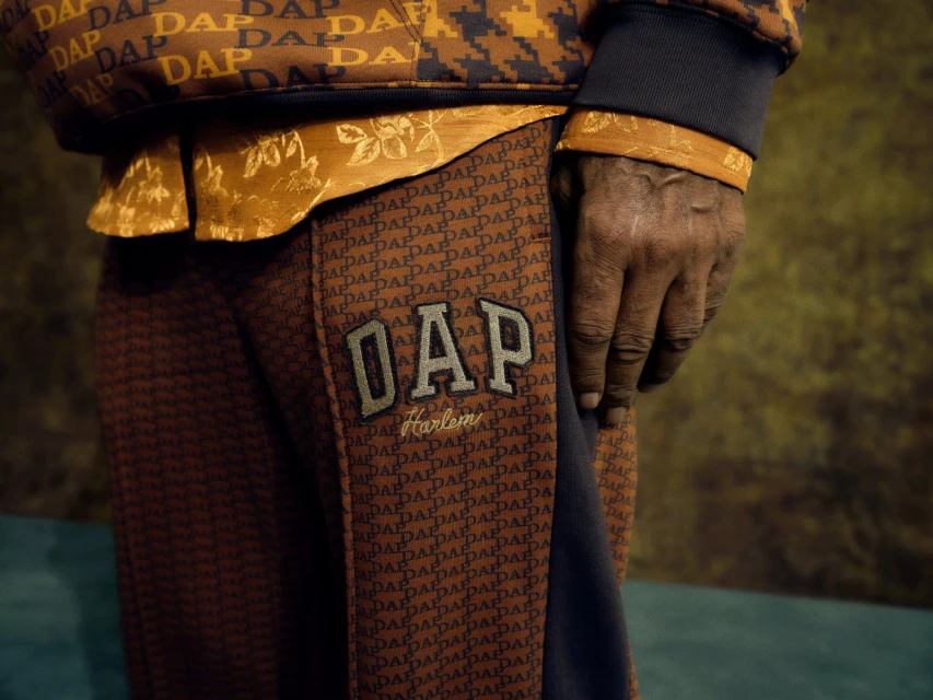 Close-up of a person's arm and torso wearing a DAP monogram patterned jacket and pants, set against a mottled background. Fashion.