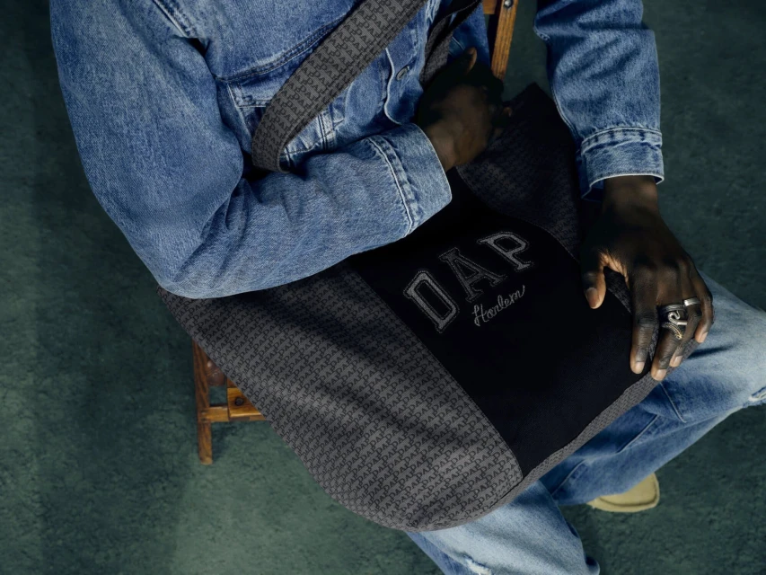 Person seated wearing a denim jacket and holding a grey shoulder bag with 'DAP Harlem' logo, showcasing urban fashion style.