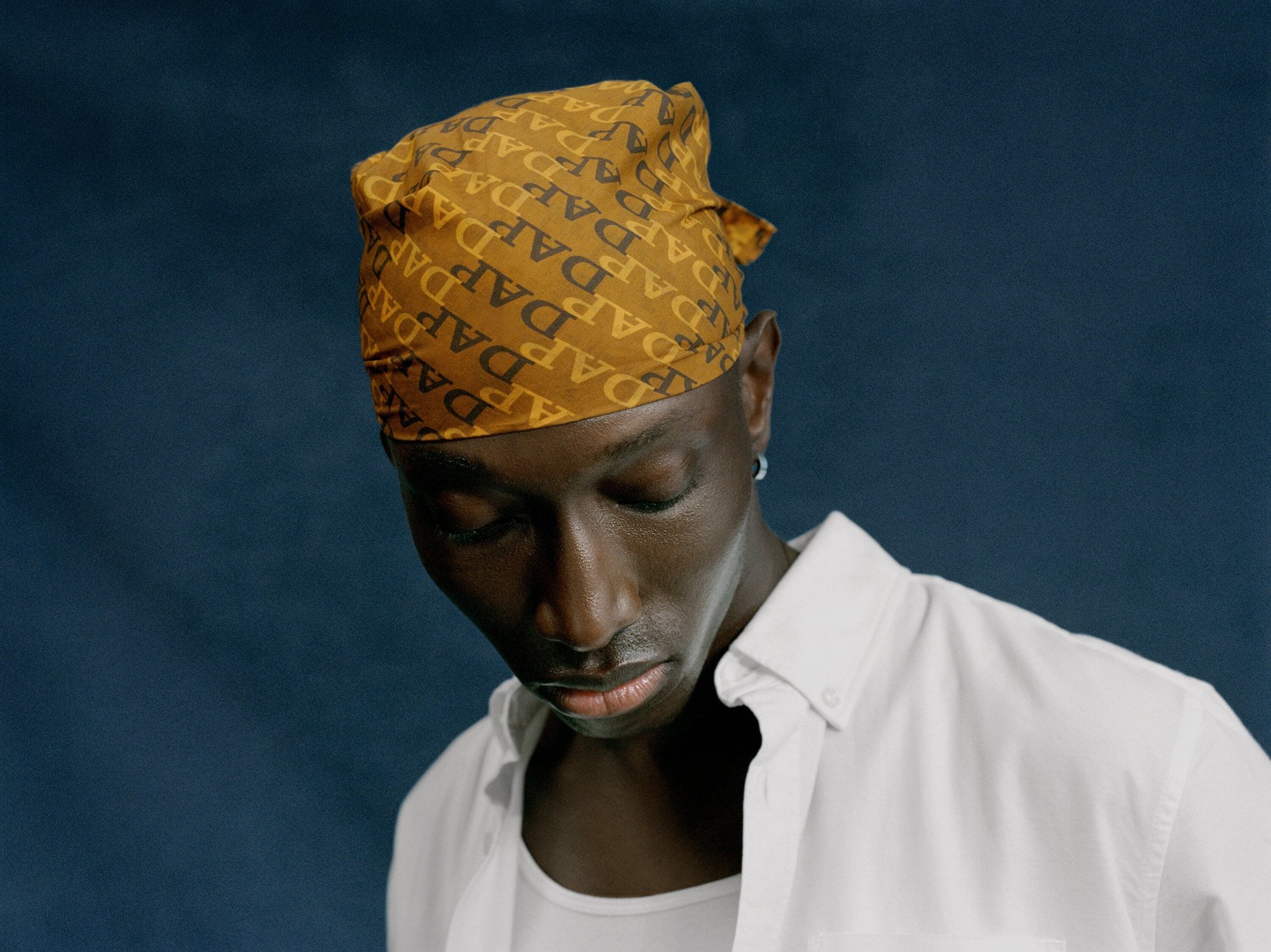 Dark-skinned man with a patterned headscarf and white button-down shirt posing against a muted blue studio backdrop with soft lighting.