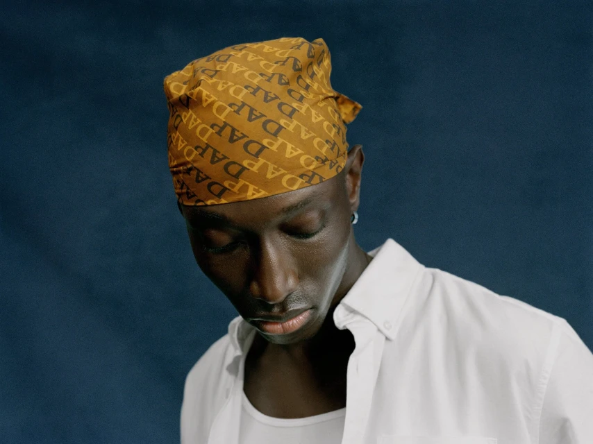 Dark-skinned man with a patterned headscarf and white button-down shirt posing against a muted blue studio backdrop with soft lighting.