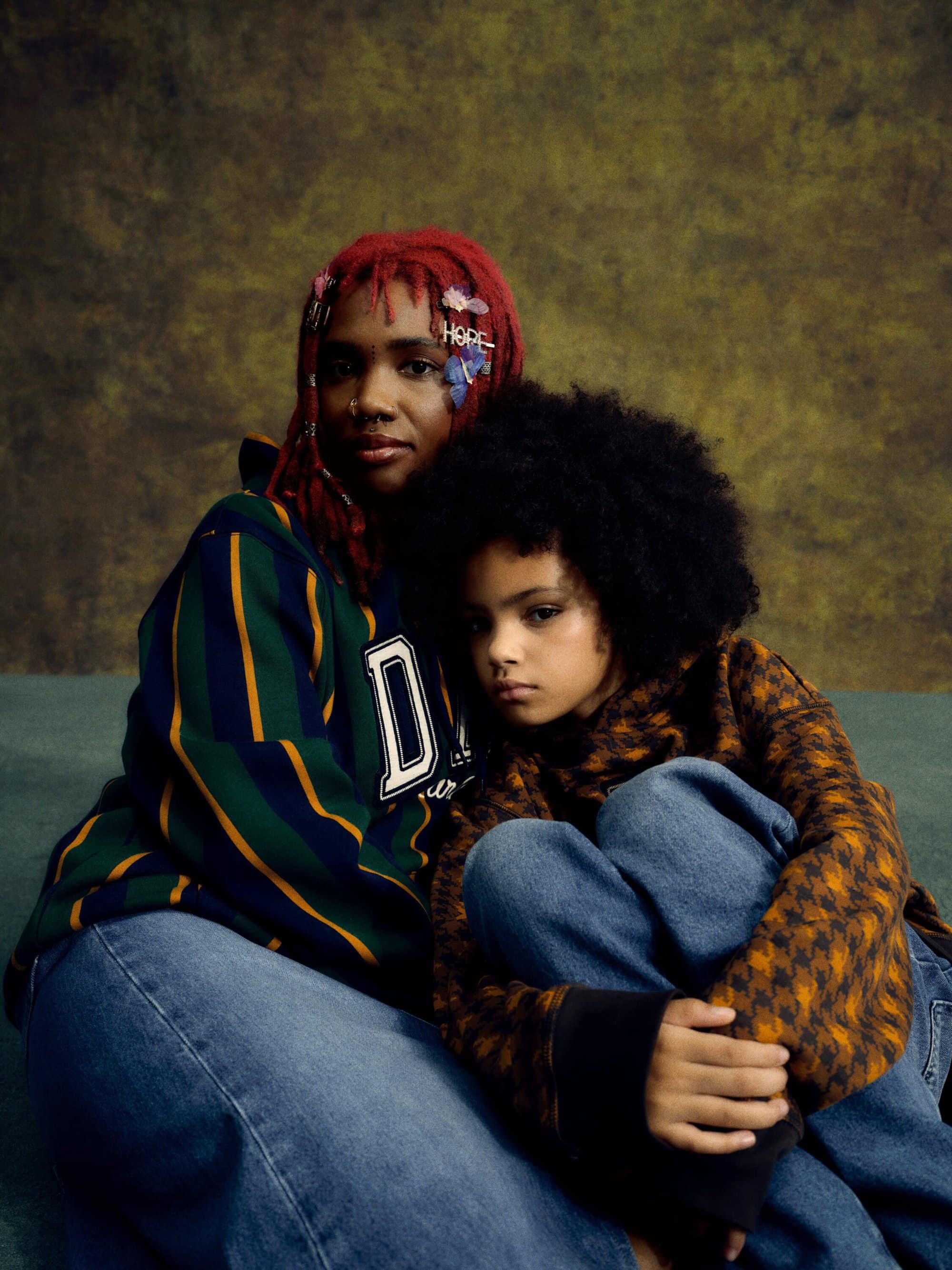 Mother and child portrait. Woman with braided red hair and hair clips sits with child with afro and patterned coat in studio setting.