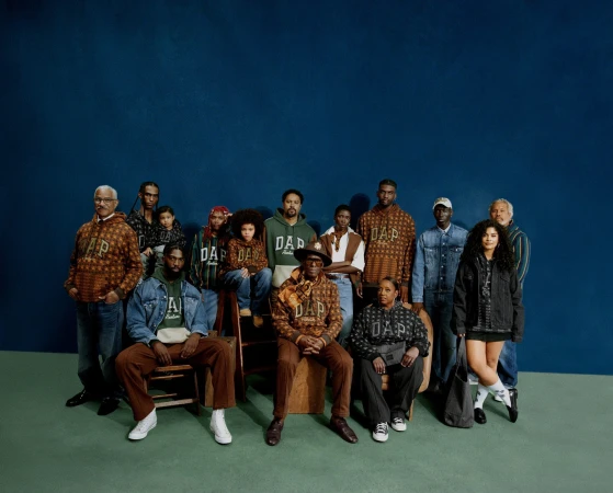 Group of people of mixed ages and ethnicities wearing GAP hoodies, posing indoors against a dark blue wall, medium shot.