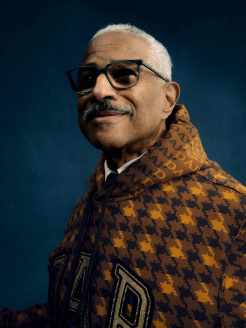 Portrait of an adult man with glasses and a mustache, wearing a patterned hoodie, set against a solid navy-blue background.