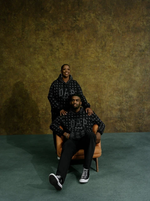 Portrait of a woman and seated man wearing matching patterned GAP hoodies and black pants against a muted gold and green background.