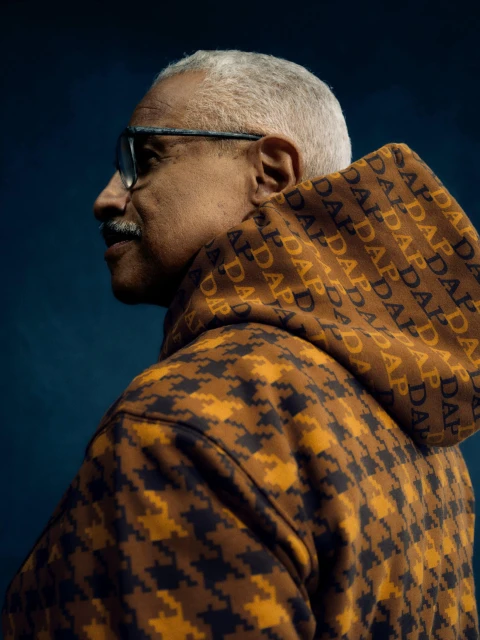 Profile shot of an adult male with grey hair, wearing glasses and a patterned hoodie, set against a dark blue background, studio lighting.