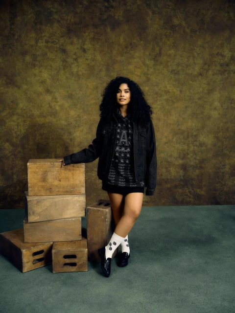 Young woman with curly hair poses in a denim jacket and patterned sweatshirt in front of wood blocks in a studio setting.