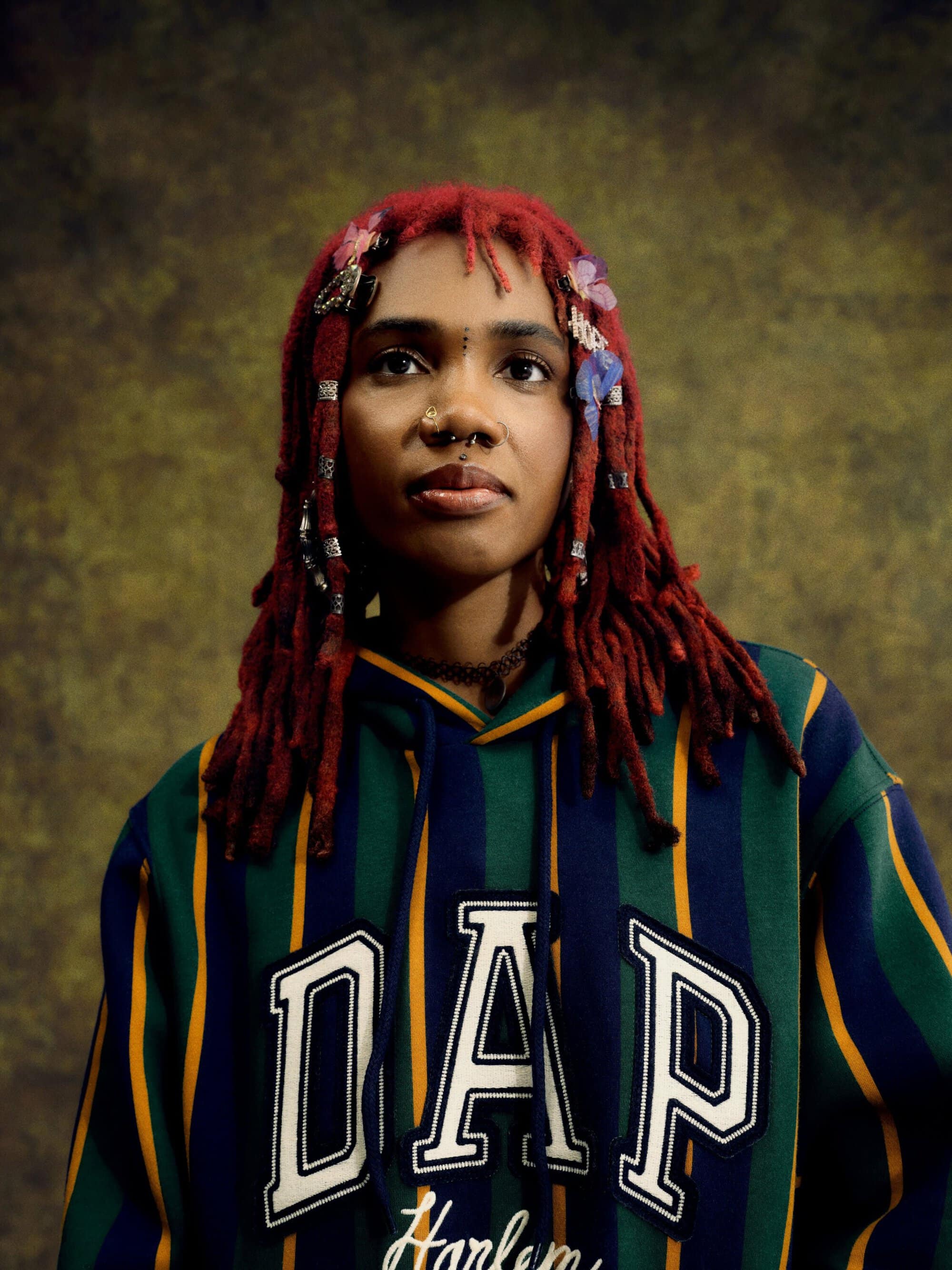 Portrait of an adult female with red dreadlocks, wearing a striped hoodie, against a mottled brown background in a studio setting.