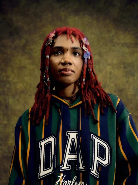 Portrait of an adult female with red dreadlocks, wearing a striped hoodie, against a mottled brown background in a studio setting.