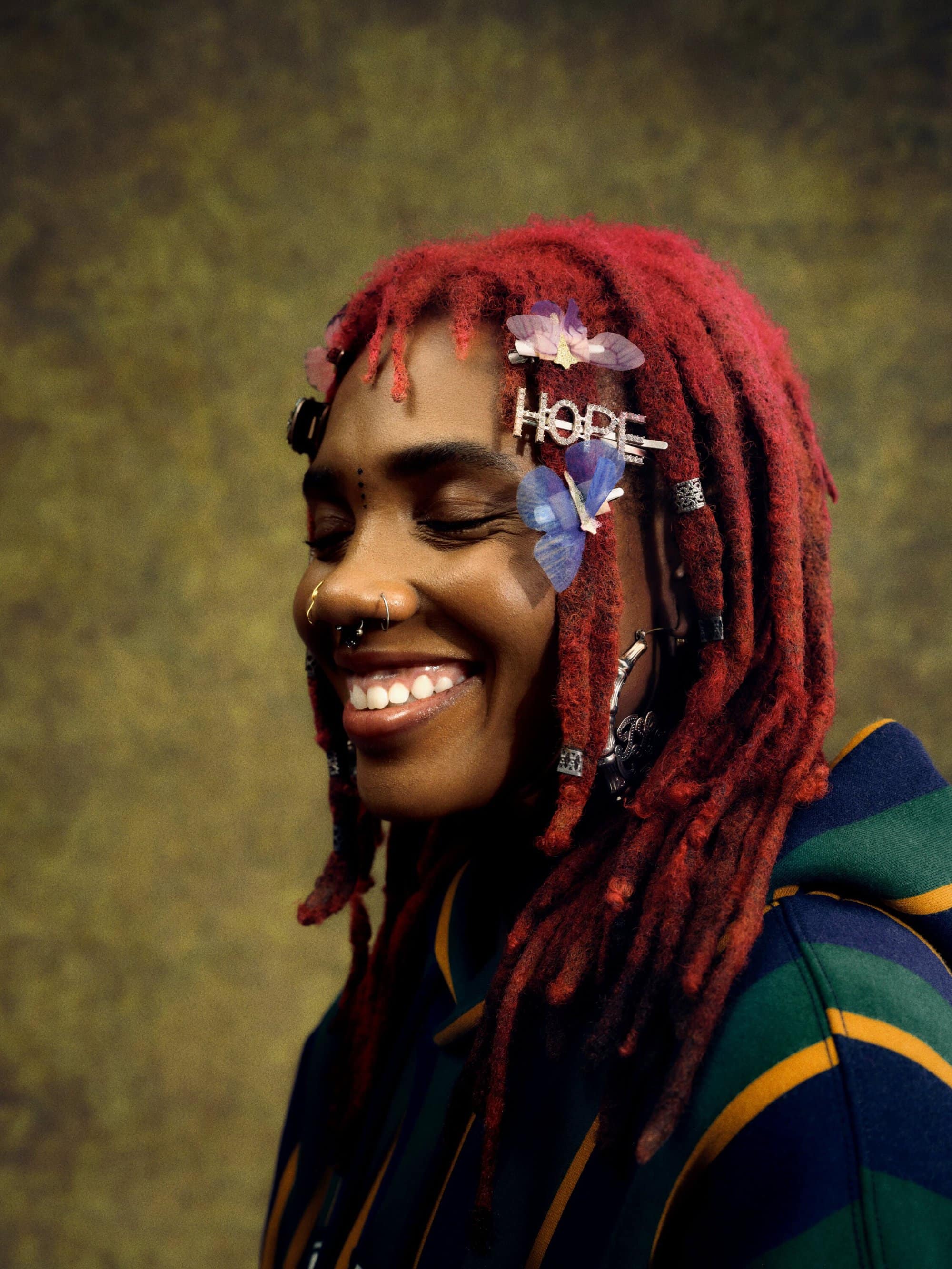 A smiling Black woman with red dreadlocks, facial piercings, and butterfly hair clips poses in a striped hoodie against a mottled backdrop.