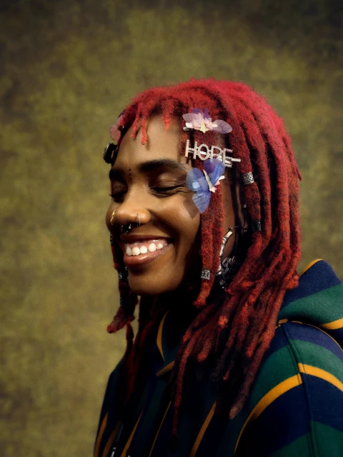 A smiling Black woman with red dreadlocks, facial piercings, and butterfly hair clips poses in a striped hoodie against a mottled backdrop.