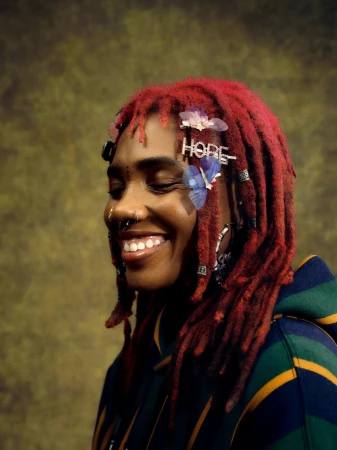 A smiling Black woman with red dreadlocks, facial piercings, and butterfly hair clips poses in a striped hoodie against a mottled backdrop.