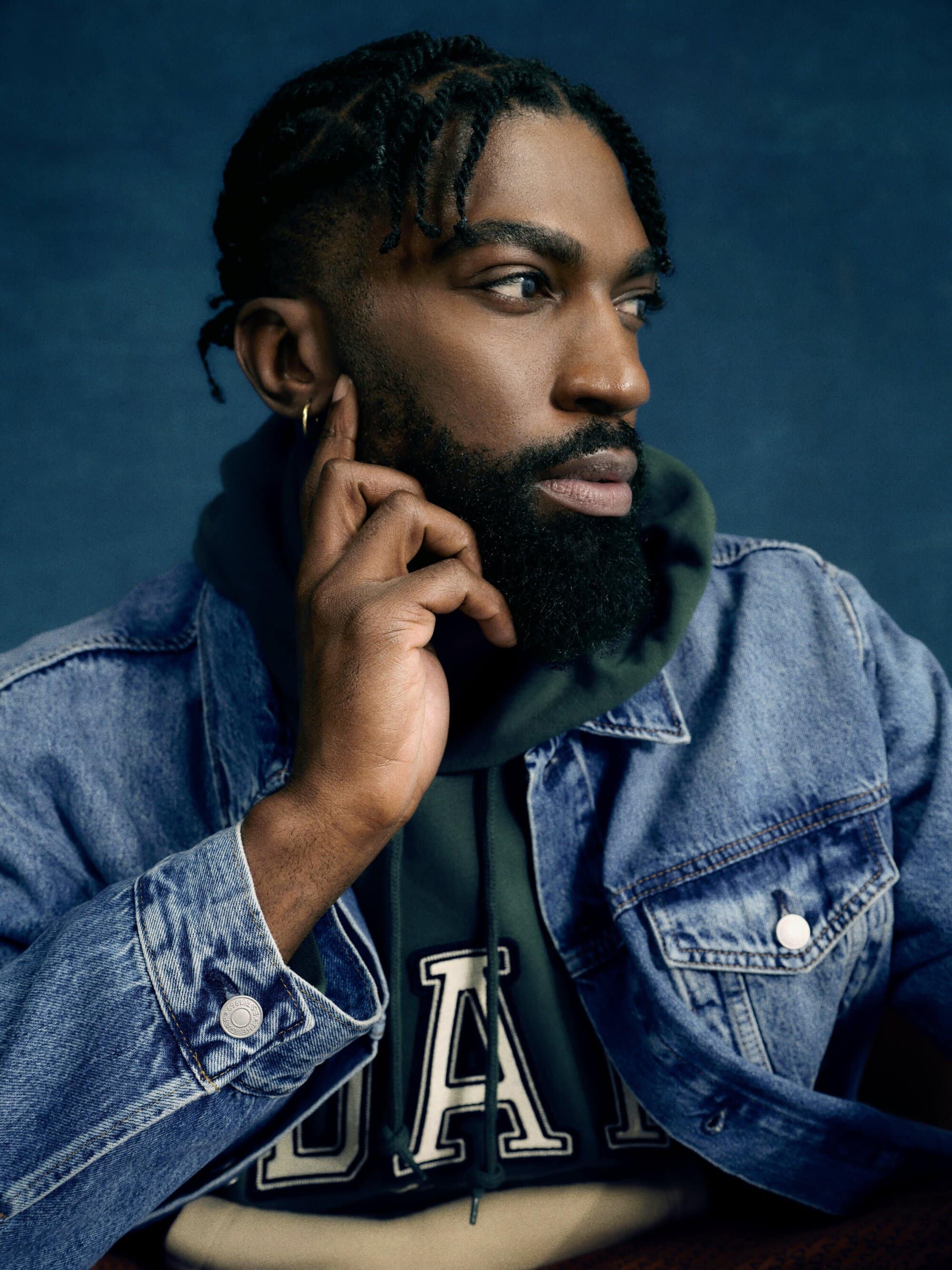 Portrait of a black man with a beard wearing a denim jacket and a hoodie with a logo, looking off to the side with a contemplative expression.