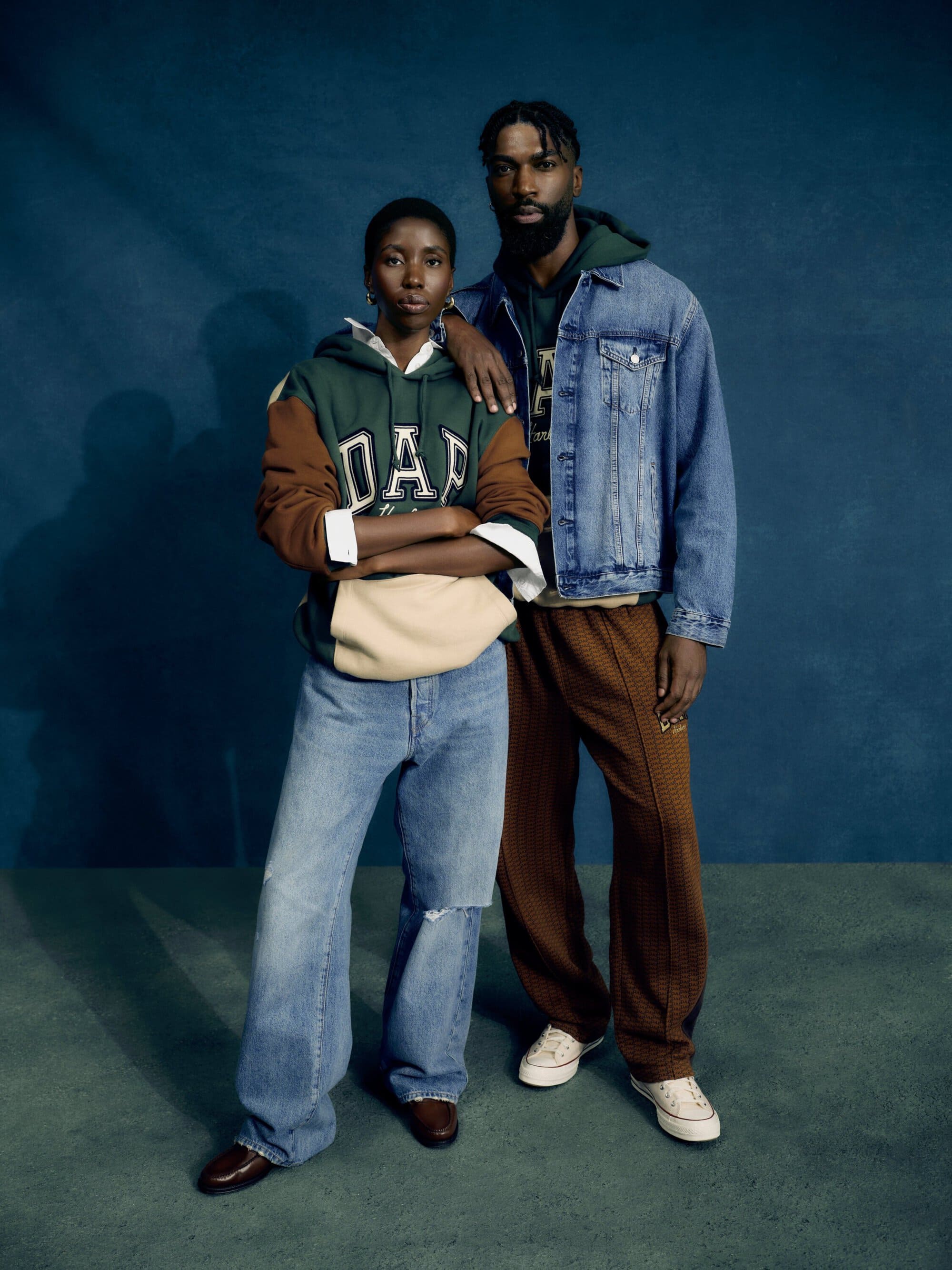 Two Black adults in casual outfits pose indoors against a blue wall. The woman wears a Gap hoodie and jeans, the man wears a denim jacket.