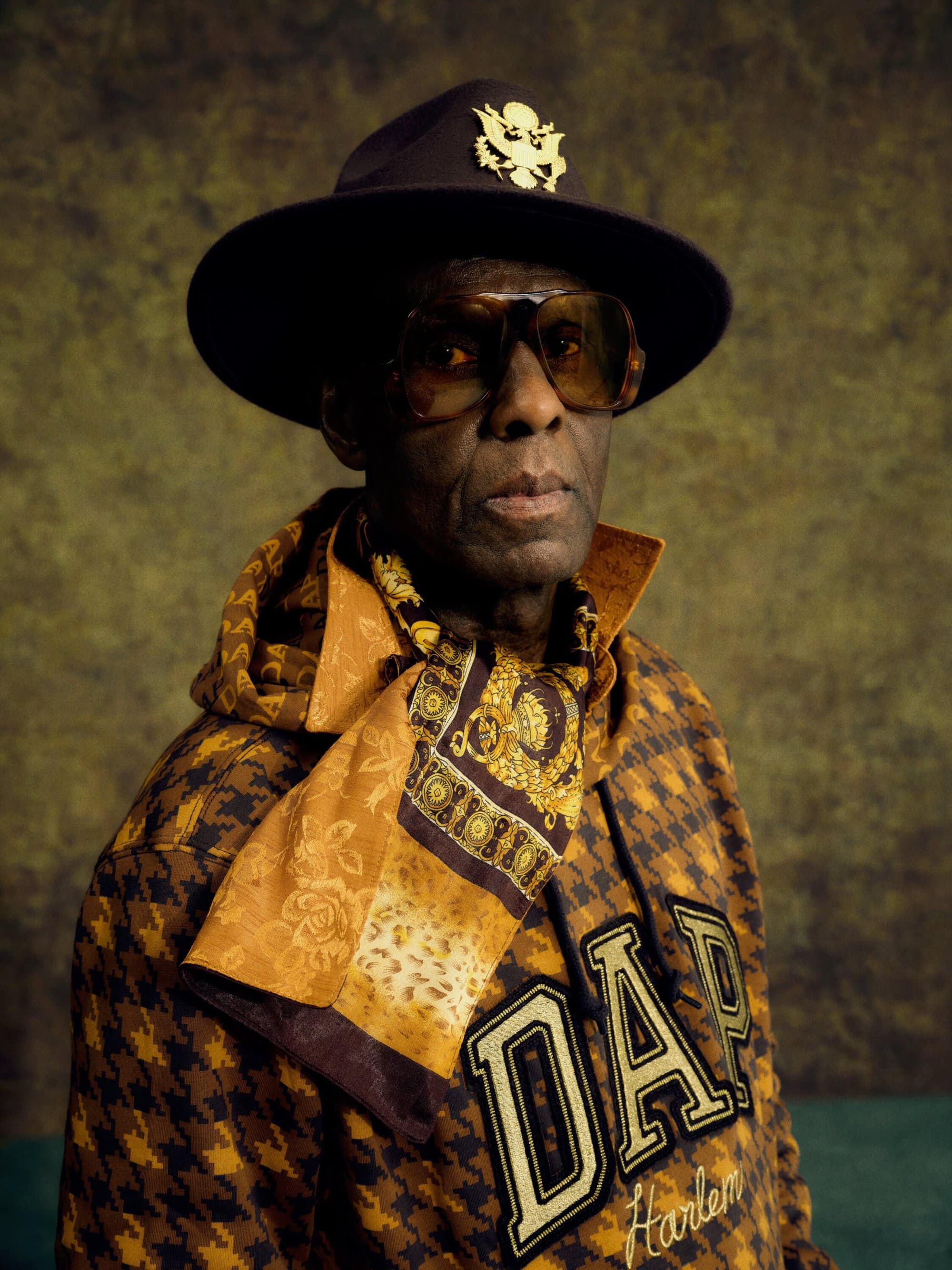 Portrait of adult male wearing a hat, sunglasses, patterned jacket, and scarf in front of a mottled yellow and green background.