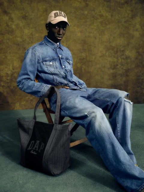 Dark-skinned male model wearing denim outfit and baseball cap poses with Gap tote bag in a studio setting with a mottled background.