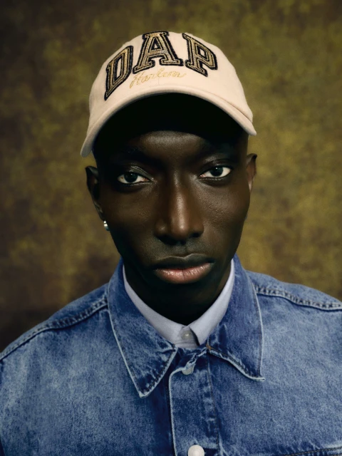 Portrait of a Black man wearing a DAP Harlem baseball cap and blue denim jacket, set against a mottled brown background. Head and shoulders.