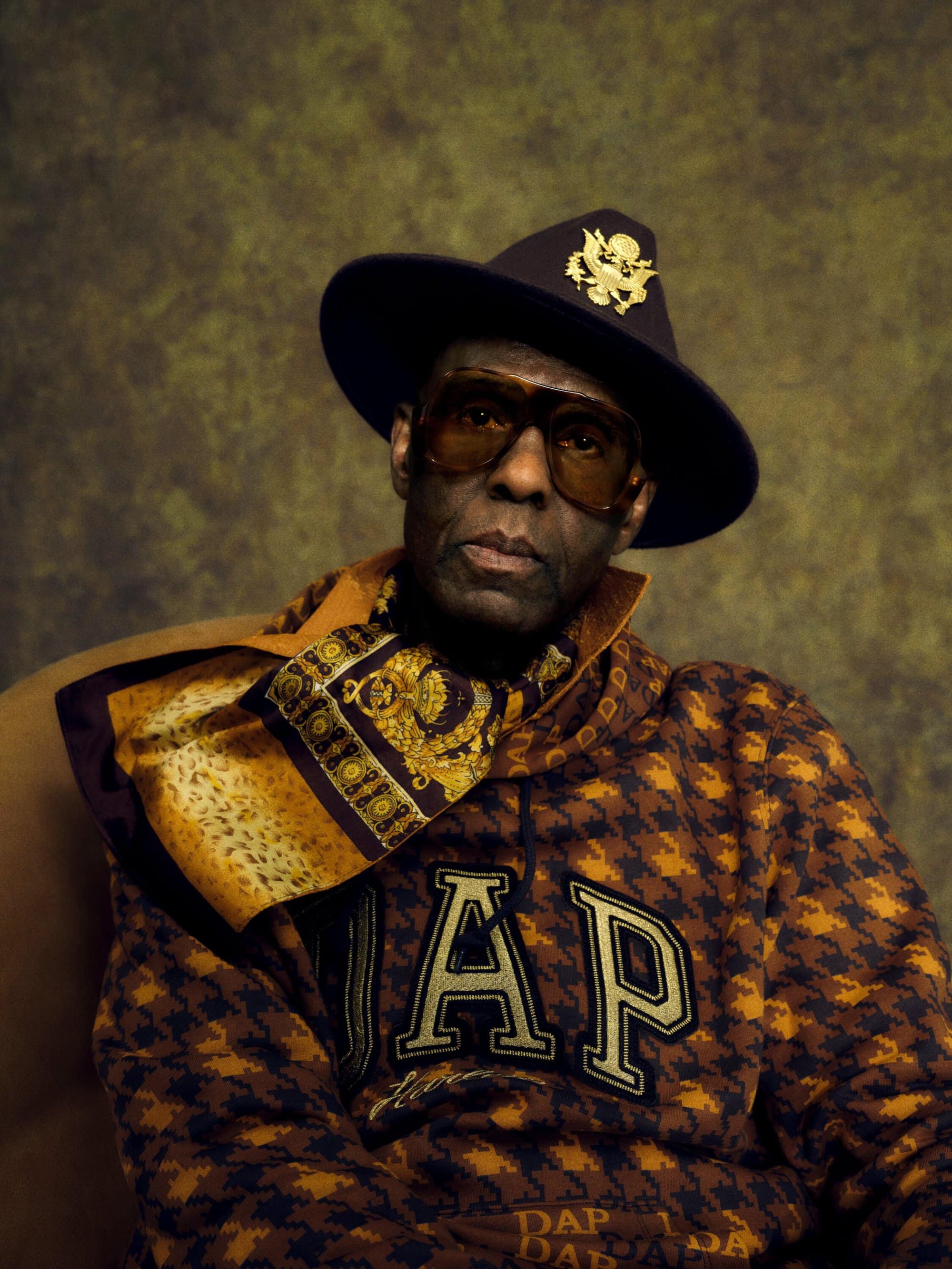 Portrait of an adult male wearing a hat and houndstooth hoodie with a scarf, against a muted golden-brown mottled backdrop.