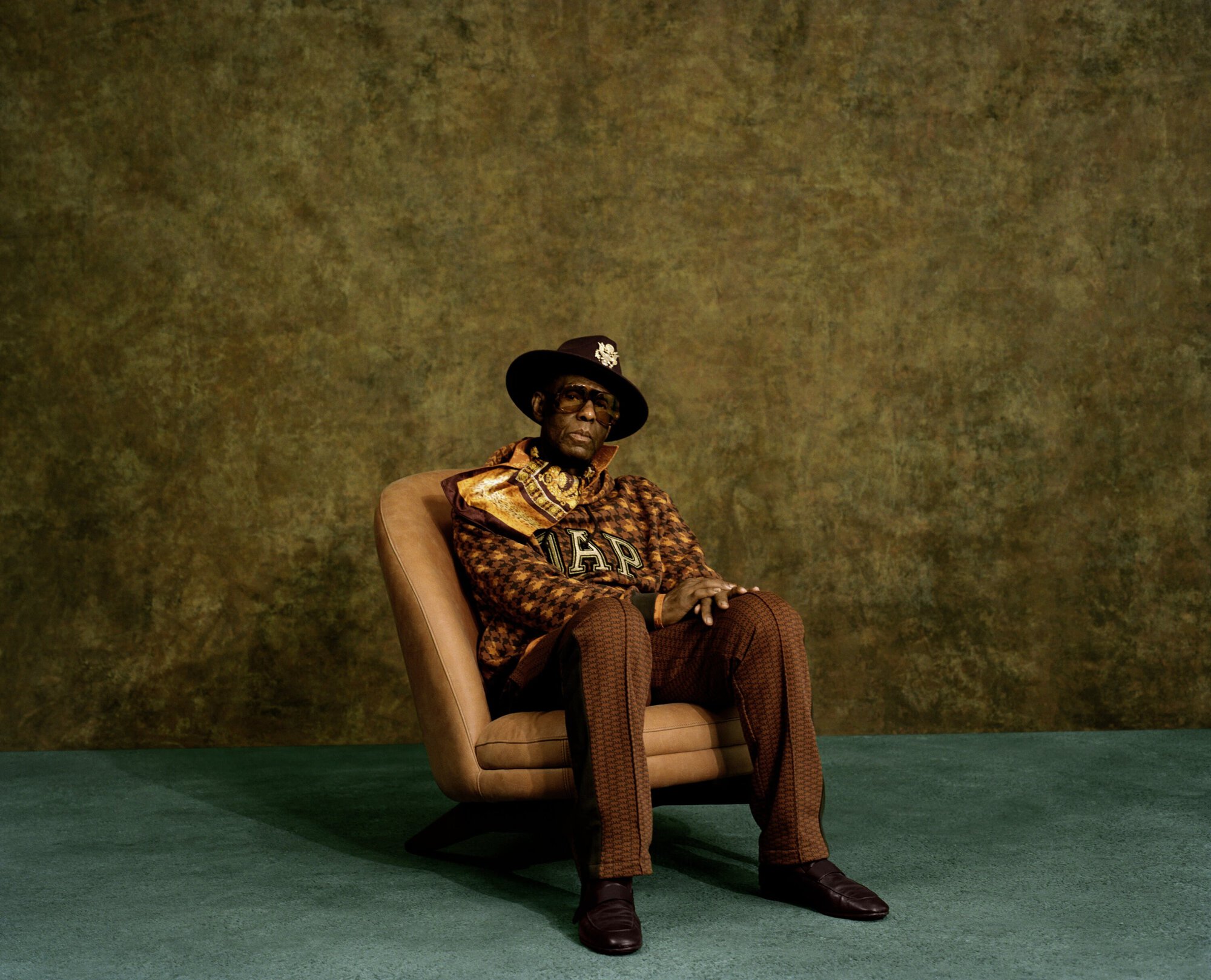 Mature African American man poses in an armchair wearing a hat, patterned jacket and scarf in front of a textured backdrop.